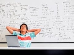 Student leaning against a whiteboard covered in equations, hands behind his head, looking overwhelmed