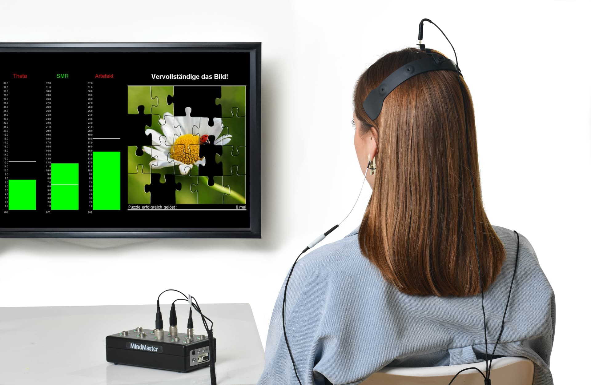 Woman with EEG headband facing a jigsaw-puzzle neurofeedback game, MindMaster device on desk