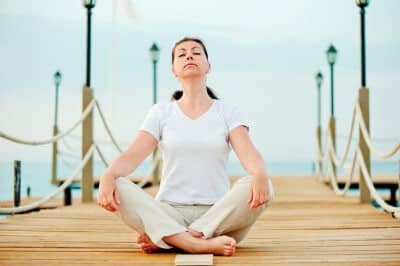 Frau sitzt mit geschlossenen Augen in Meditationshaltung auf einem Holzsteg